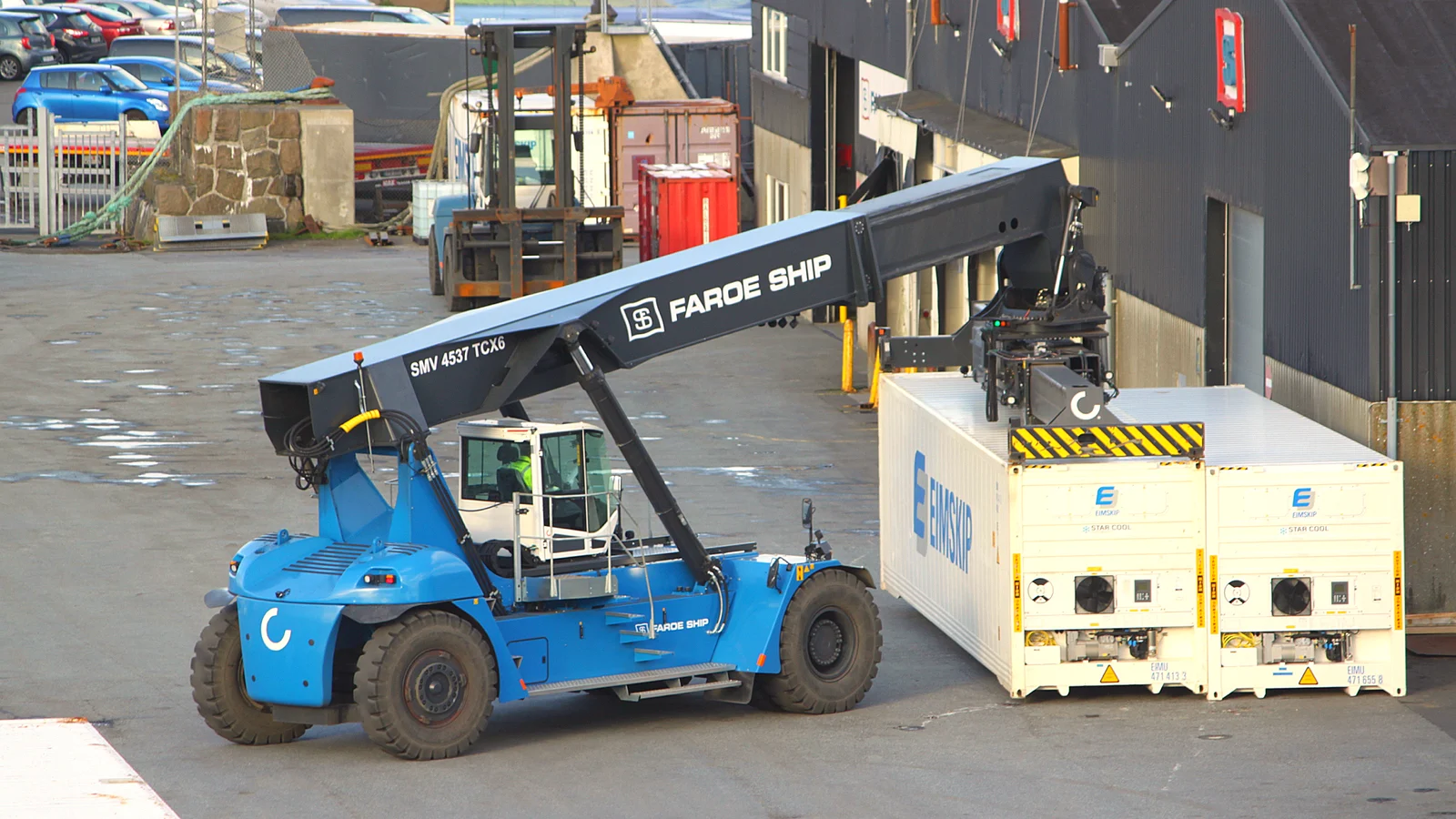 Blue reach stacker beside two white refrigerated containers in a port yard