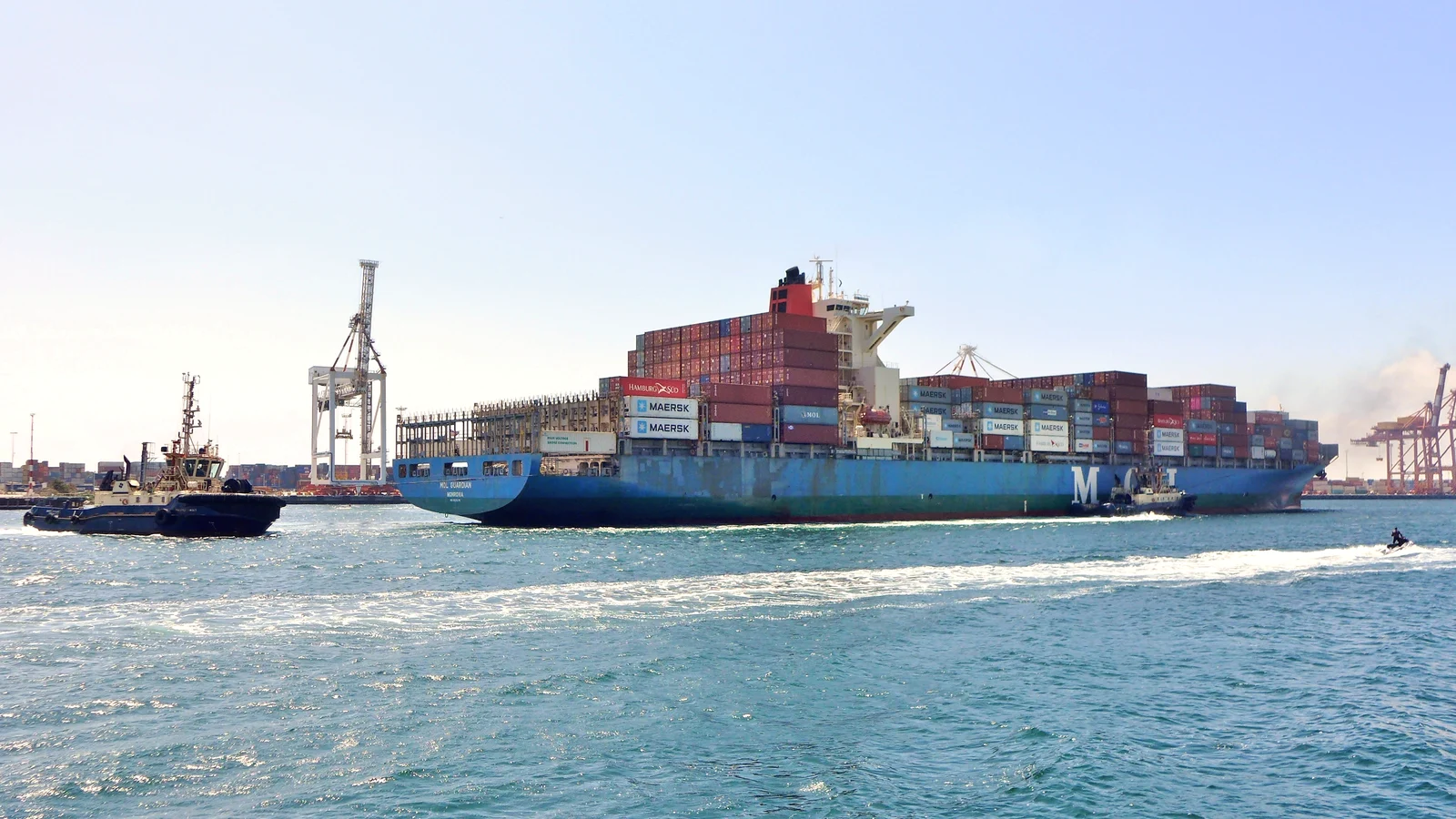 Loaded container ship escorted by a tug arriving in harbour under blue sky