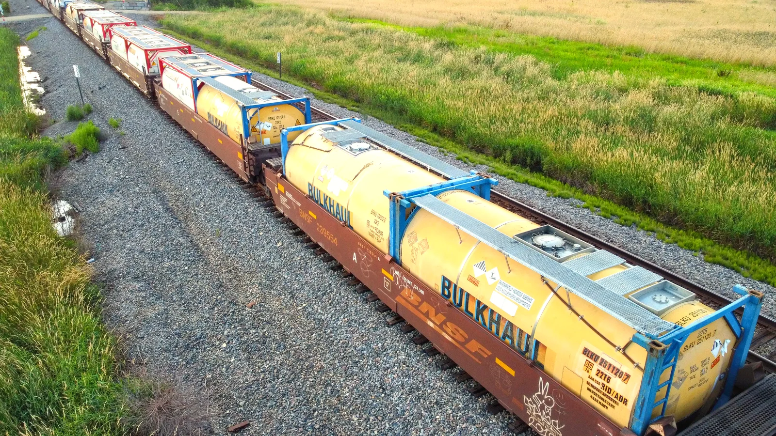 Freight train carrying yellow and blue bulk tank containers past green fields