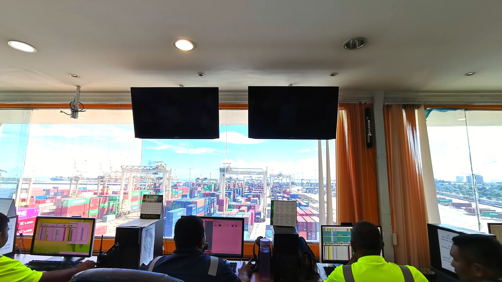 Port control room with monitors overlooking a container terminal with cranes and stacked boxes