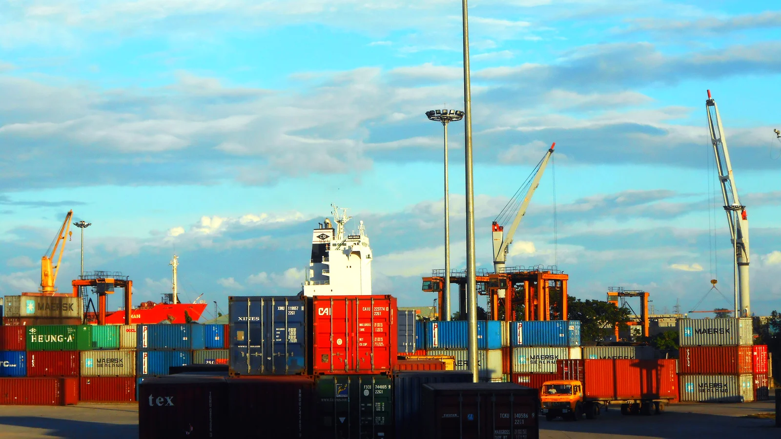 Stacks of shipping containers in a port yard under blue sky with cranes