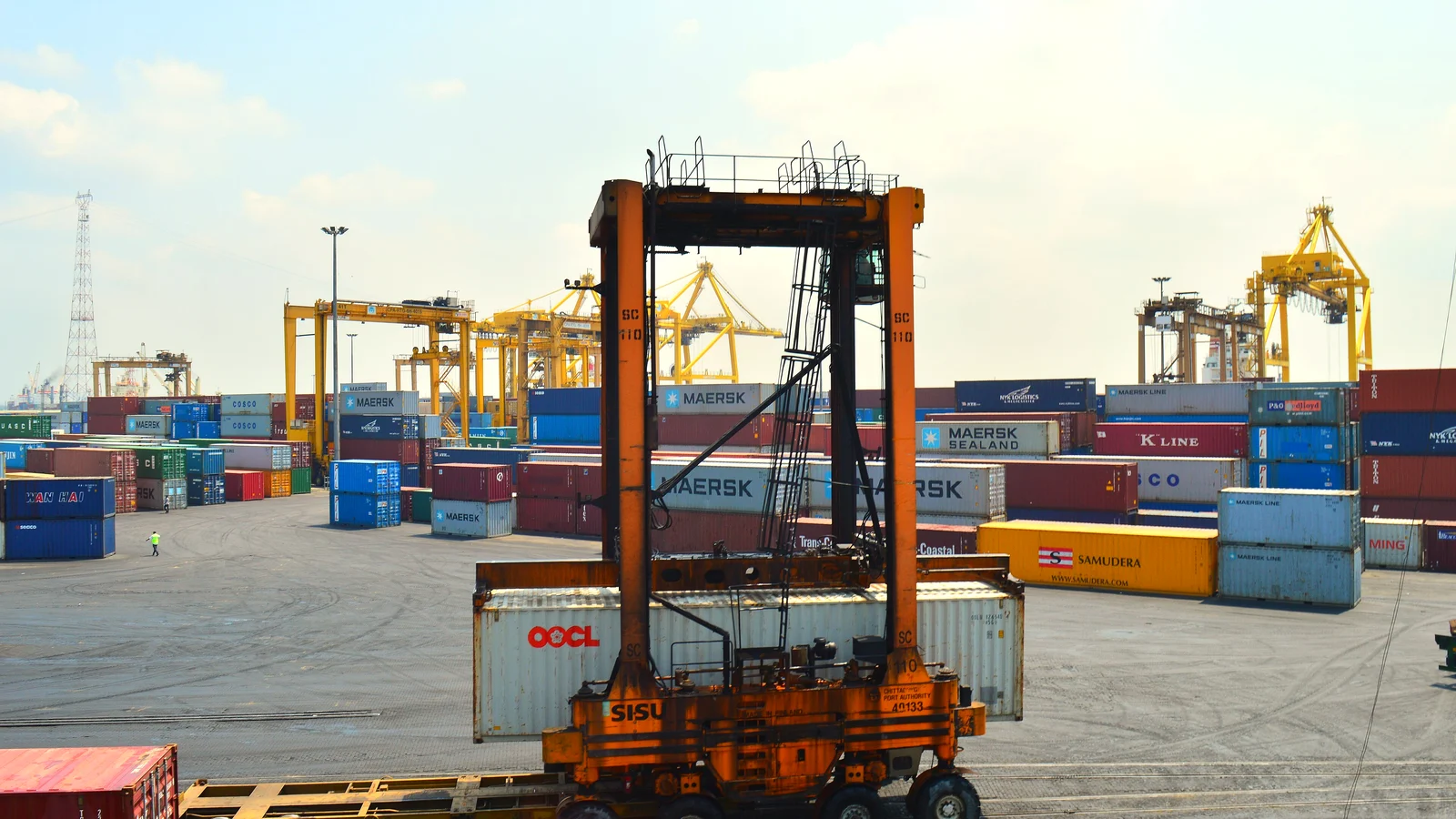 Straddle carrier lifting a container in a terminal yard with colourful container stacks and cranes